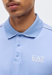 Άνδρας που φοράει ανοιχτό γαλάζιο πόλο EA7 Emporio Armani με ριμπ ύφασμα στο γιακά και τρία κουμπιά κουμπωμένα, με τα χείλη και την κάτω γνάθο μερικώς ορατά.