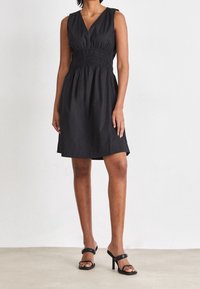 Svart ärmlös klänning med V-halsringning, rynkad midja och utsvängd kjol. Slät, lättvikts tyg med mjuk textur, bärs med svarta klacksandaler.