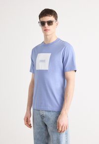 T-shirt de algodão azul claro com mangas curtas, com um design de logotipo quadrado branco. Usado com jeans de ganga azul claro e óculos de sol transparentes.