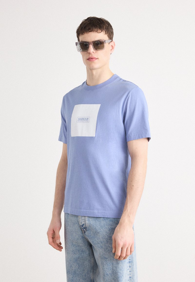 T-shirt de algodão azul claro com mangas curtas, com um design de logotipo quadrado branco. Usado com jeans de ganga azul claro e óculos de sol transparentes.