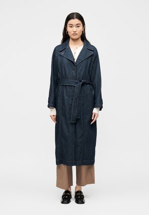 Femeie stând cu fața înainte, purtând o trenci din denim închis la culoare cu curea, pantaloni bej cu picioare largi, top alb, mocasini negri și cercei de aur în cerc.