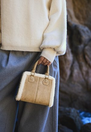 Osoba ubrana w kremowy sweter i szare spodnie, trzymająca małą beżową torebkę z białym futrzanym wykończeniem za uchwyty.