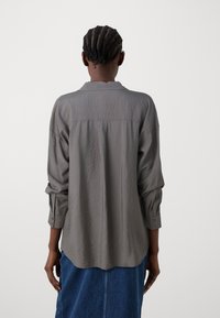 Blusa cinza de corte solto, com um tecido texturizado e amassado, mangas longas, punhos com botões e uma bainha arredondada, exibida pela parte de trás.