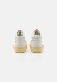 Bijele visoke tenisice od teksturirane kože, s gumenim žutim potplatom i podstavljenim ovratnikom. Nema vidljivih logotipa ili ukrasa.