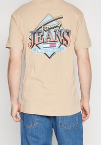 Béžové bavlněné tričko s barevným potiskem na zádech, na kterém je nápis "Tommy Jeans" a "New York City" uvnitř diamantového tvaru.