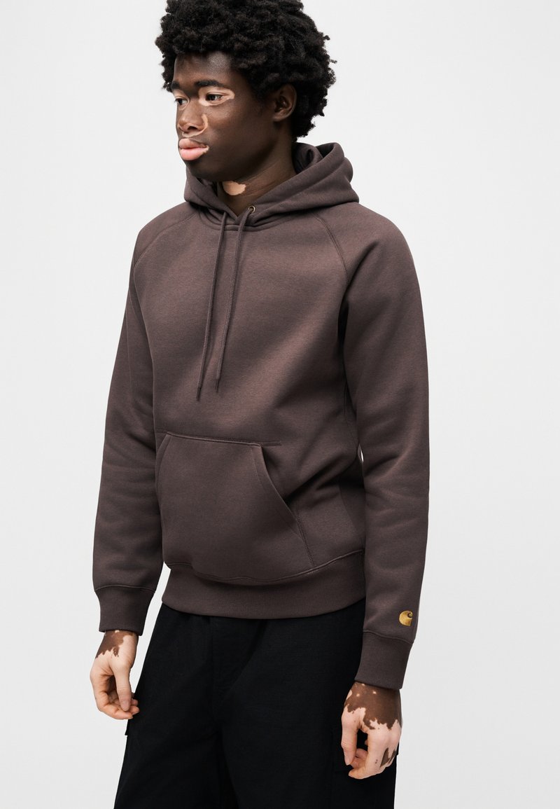 Brun hættetrøje med kengurulomme, snøre i hætten og ribbede manchetter. Har en logopatch på venstre ærme. Blødt, tekstureret stof.