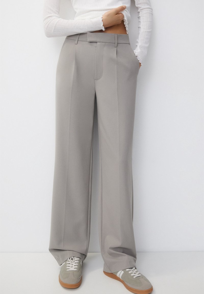 Pantalons à jambe large en tissu gris clair avec une texture lisse, présentant des plis à l'avant et une coupe ajustée, associés à des sneakers grises.