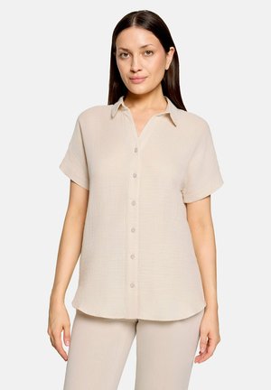 Kvinde iført beige kortærmet skjorte med knapper og matchende lyse bukser, stående mod en ensfarvet lys baggrund.