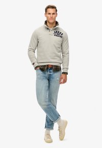 Lichtgrijze sweatshirt met een kraag en marineblauw print. Gedragen over een donker geruite shirt. Gecombineerd met lichtblauwe spijkerbroek en beige sneakers.