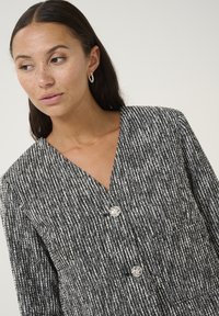 Kobieta z długimi ciemnymi włosami i piegami, nosząca srebrne kolczyki koła oraz czarno-białą, teksturowaną kurtkę z guzikami, patrząca w lewo.