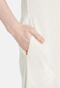 Ivory linen dress featuring a side pocket, with a textured, lightweight fabric and subtle woven pattern. Hand resting in the pocket.