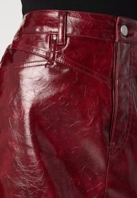 Gros plan sur une jupe en cuir rouge profond brillante avec couture visible, passants de ceinture et fermeture par bouton métallique.