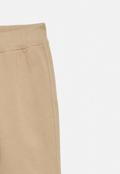 Közeli kép bézs kötött anyagú nadrágról, amely rugalmas derékpántot és oldalsó varrást mutat egyszerű háttéren.