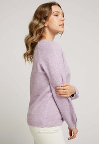Pull en tricot lilas à manches longues, avec une coupe décontractée et un ourlet côtelé. Associé à un pantalon blanc, montrant une texture douce et une couleur subtile.