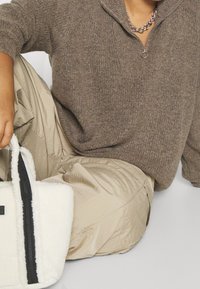 Pull en tricot marron avec une fermeture éclair au quart et des manches longues, associé à un jogging beige brillant. Sac blanc avec une fermeture éclair noire et des accents texturés.