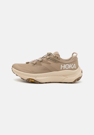 Beige kalnų vaikščiojimo batai su tekstūruotu paviršiumi, Vibram padu ir elastinėmis raištelėmis. Yra HOKA logotipas ir GORE-TEX ženklas, užtikrinantis vandens atsparumą.