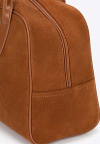 Bruine suède handtas met een gestructureerde vorm, voorzien van een ritssluiting en gestikte details langs de zijkanten voor duurzaamheid.