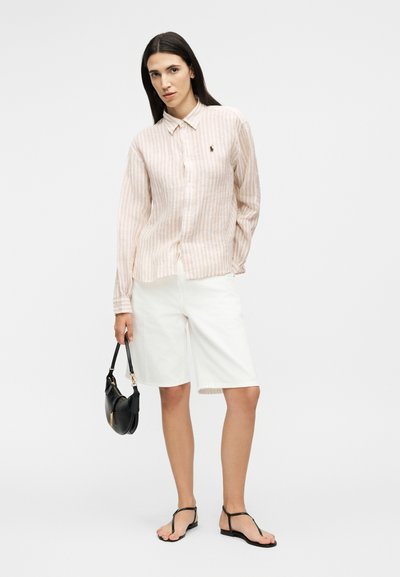 Femme portant une chemise beige à rayures à manches longues avec boutons, un short blanc à hauteur des genoux, des sandales noires, tenant un petit sac à main noir.