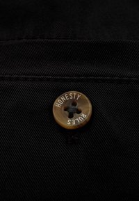 Schwarzer Stoff mit diagonaler Struktur, ausgestattet mit einem braunen Knopf, der mit "EHRLICHKEIT REGIERT" beschriftet ist und ein klassisches Vierlochdesign aufweist.