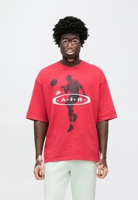 Κόκκινο φαρδύ βαμβακερό t-shirt με μαύρο γράφημα καλαθοσφαιριστή, λευκό οβάλ σχήμα και το κείμενο "Air". Το μοντέλο φοράει ανοιχτό πράσινο παντελόνι.