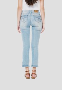 Lichtblauwe denim jeans met een slim-fit, gestikte zakken en een leren merklabel aan de achterkant. Draagt bruine sandalen met bandjes.