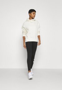 Felpa in morbido tessuto crema con tasca a marsupio e coulisse regolabile. Leggings neri con logo bianco di lato. Sneakers bianche.