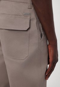 Ciemnoskóra dłoń spoczywająca obok, ubrana w taupe'owe spodnie Armani Exchange z widoczną tylną kieszenią i metalową metką marki.