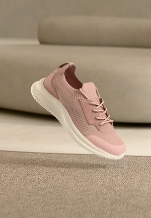 Pantof sport tricot roz deschis cu talpă albă, șireturi în față și călcâi căptușit, așezat lângă o canapea bej pe un covor cu textură.