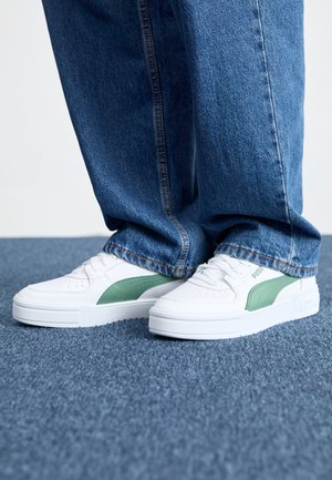 Persona que lleva zapatillas blancas Puma con rayas verdes y pantalones vaqueros anchos azules de pie sobre un suelo alfombrado azul.