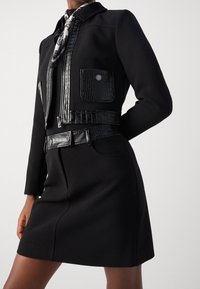 Veste noire avec finitions en similicuir, fermeture éclair à l'avant, deux poches plaquées et ceinture ajustée. Accompagnée d'une écharpe à motifs.