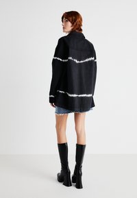 Jachetă din denim negru cu model alb tie-dye, croi lejer, cu guler, purtată cu o fustă din denim și cizme negre cu toc până la genunchi.