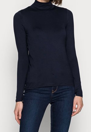 Personne portant un haut noir ajusté à col roulé à manches longues et un jean bleu foncé, debout devant un fond clair uni.