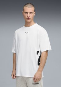 T-shirt blanc à manches courtes avec logo Puma noir sur la poitrine, présentant un motif géométrique noir sur le côté et une coupe ample.