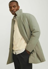 Abrigo acolchado verde con cuello alto, con cierre de cremallera. Llevado sobre una blusa crema con dobladillo a rayas y pantalones negros.