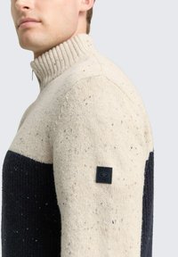 Mann trägt einen hoch geschlossenen, gesprenkelten Pullover in Creme und Schwarz mit einem kleinen schwarzen Patch auf dem oberen Ärmel.