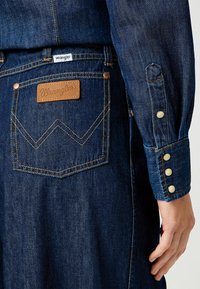 Takapocket tummansinisissä Wrangler-farkuissa, joissa on erivärinen ommel, nahkainen brändilogo, ja käsi lepää denimitakin hihassa, jossa on napit.