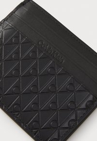 Porte-cartes noir en cuir texturé, présentant un motif géométrique et le logo "Calvin Klein" embossé sur la surface.