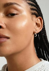 Orecchino a cerchio in argento con design a forma di luna crescente, arricchito da piccole pietre trasparenti. La superficie texturizzata riflette la luce. Capelli intrecciati in vista.