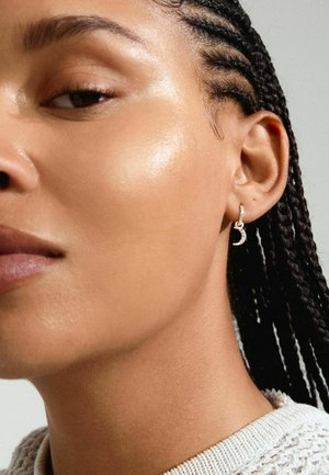 Silver hoop earring featuring a crescent moon design, set with small clear stones. Textured surface reflects light. Braided hair visible.