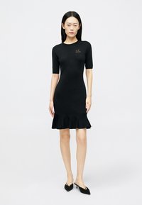 Μαύρο εφαρμοστό φόρεμα με κοντά μανίκια, ριμπ υφή και λεπτομέρεια βολάν στο τελείωμα. Διαθέτει μικρό κίτρινο λογότυπο στο στήθος. Ζευγάρι μαύρα παπούτσια.