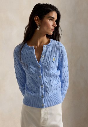 Mujer con cabello largo y oscuro, lleva pendientes plateados y un cárdigan abotonado de punto trenzado azul claro combinado con pantalones de color claro.