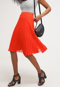 Kvinna i en röd plisserad kjol, svarta klacksandaler och en vit randig topp, som håller en svart axelväska.