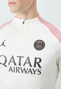 Valkoinen urheilu paita, jossa on pinkit olkapääkorostukset, vetskari kaulassa, musta "QATAR AIRWAYS" -teksti ja pyöreä "PARIS SAINT-GERMAIN" -merkki.