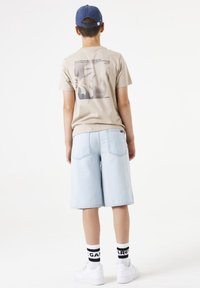 T-shirt graphique beige avec un imprimé sombre, associé à un short en denim bleu clair. Des baskets blanches et des chaussettes à rayures noires complètent la tenue.