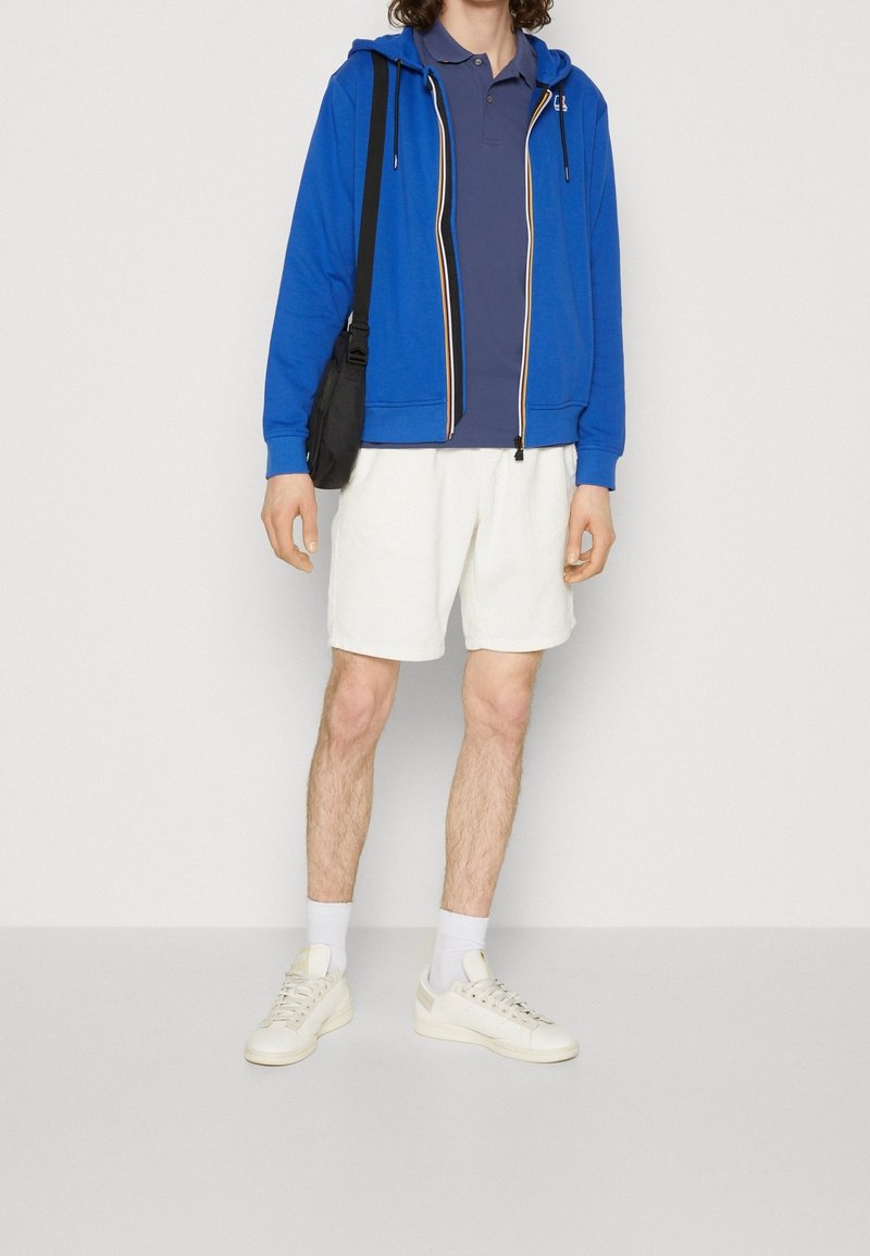 Personne portant un sweat à capuche zippé bleu sur un polo bleu marine, un short blanc, des baskets blanches et portant un sac bandoulière noir, debout devant un fond uni.