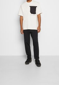 Bílá tričko s černou kapsou na zip a grafickými detaily, doplněné černými sportovními kalhotami a černými teniskami.