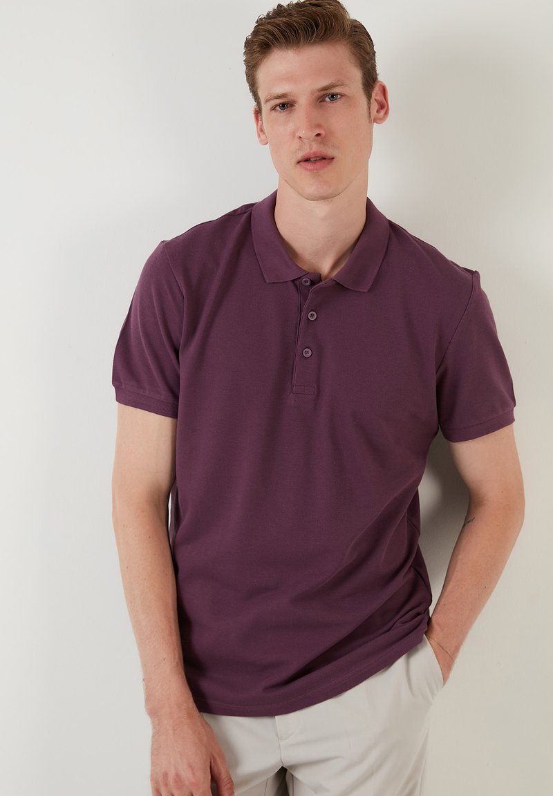 Vijolična polo majica s ovratnikom, zapenjanjem na tri gumbe, kratkimi rokavi in gladko teksturo tkanine. Model pozira z rokama v žepih.