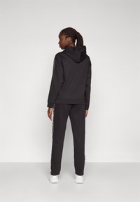 Schwarzer Hoodie mit einer Kordelzugkapuze und gerippten Bündchen, kombiniert mit passenden Jogginghosen mit seitlichem Logo-Detail, getragen mit weißen Sneakern.
