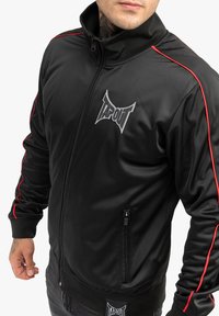 Chaqueta de chándal negra con cremallera y franjas laterales rojas, que presenta un cuello alto, puños ajustados y un prominente logo plateado "TAPOUT" en el pecho.