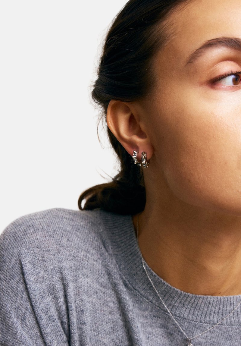 Sidabrinės auskarai-žiedai su tekstūruotais trikampių akcentais. Modelis vilki pilką megztinį, kuris aiškiai parodo auskarus prieš jos odą.